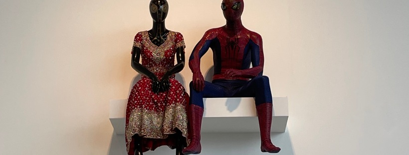 A spiderman manequin sits on a floating shelf next to another manequin in traditional indian dress